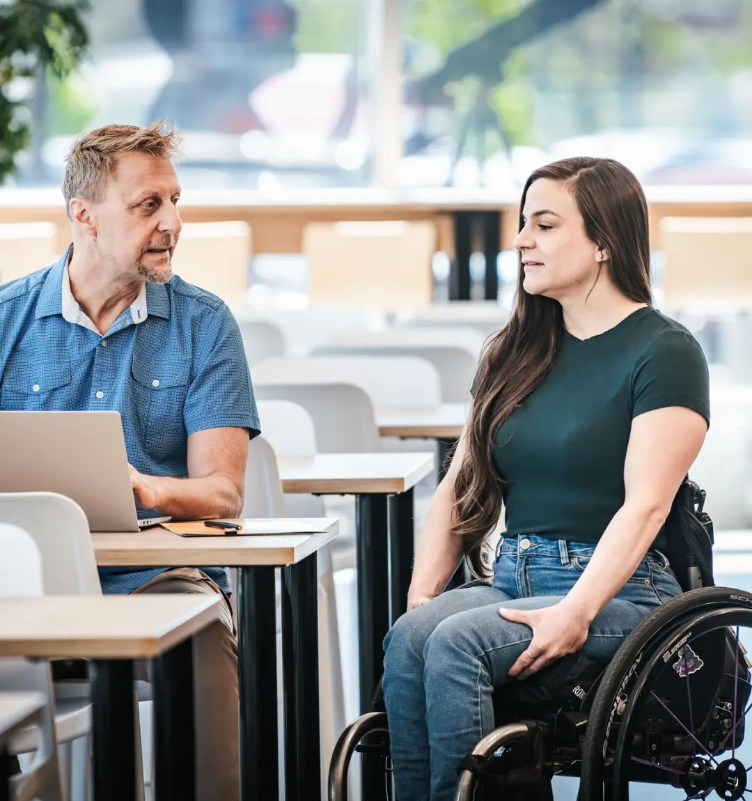 Une femme en fauteuil roulant échange avec un homme assis devant un ordinateur portable.