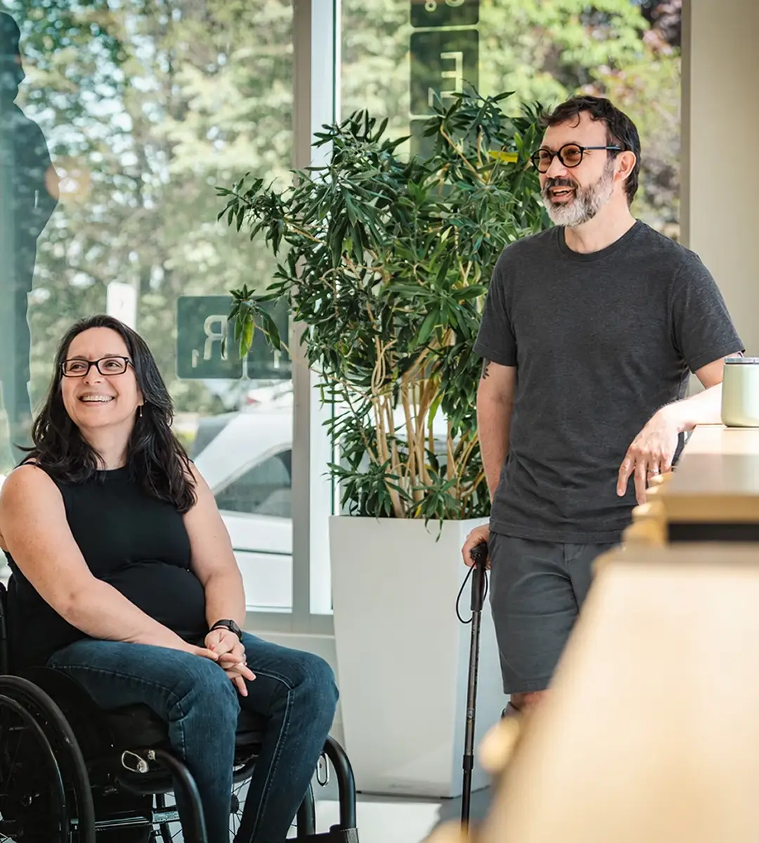 Une femme en fauteuil roulant et un homme debout avec une canne sourient dans un espace lumineux.